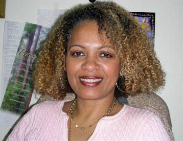 A woman with curly hair, wearing a pink top and a necklace, smiling at the camera.