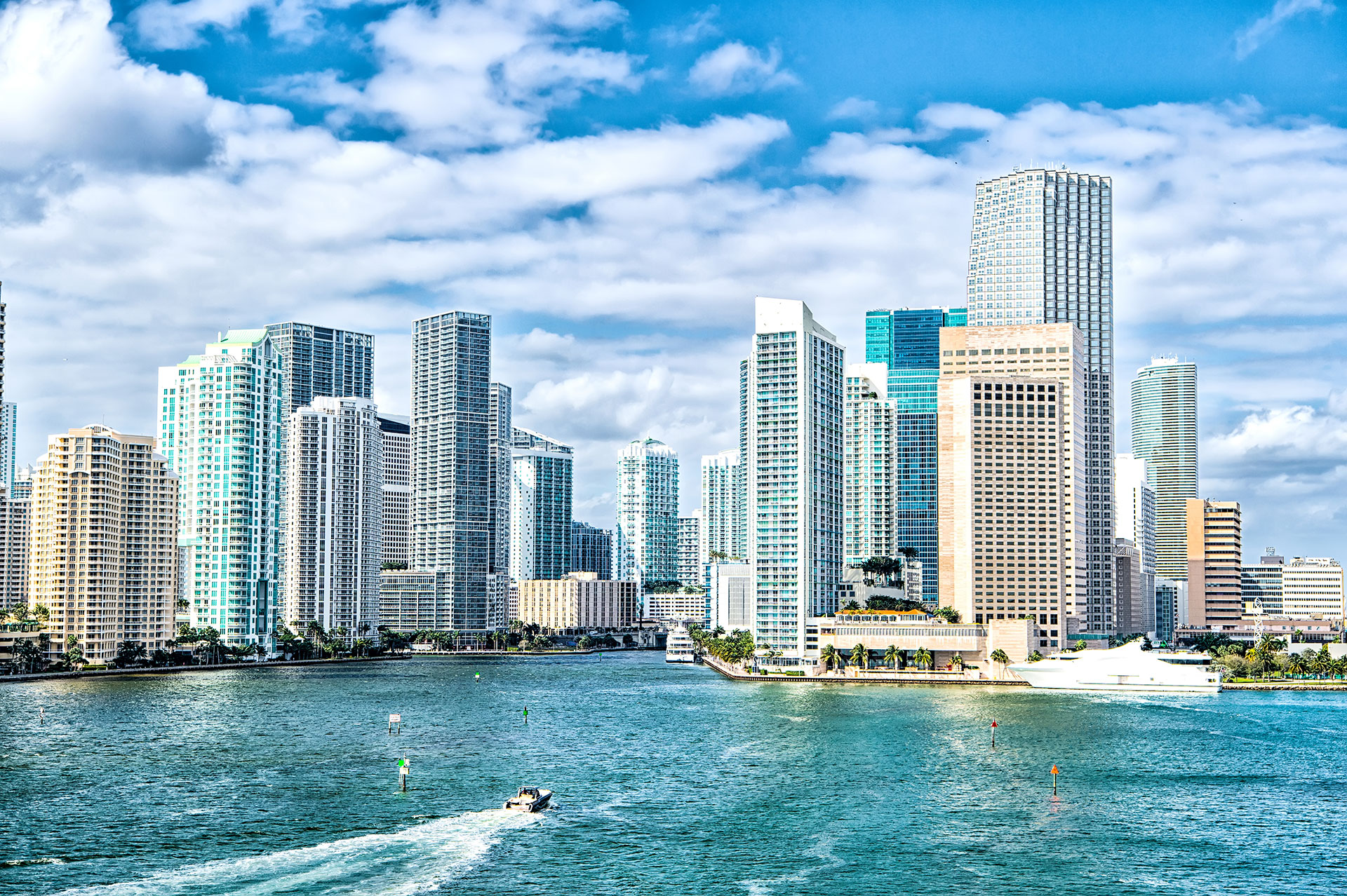 Aerial view of a city skyline with high-rise buildings, a river, and boats, under a clear blue sky.