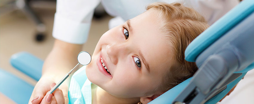 A young child is seated in a dental chair, smiling and holding a toothbrush, with a dentist in the background.