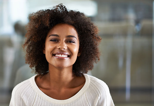 A smiling woman with curly hair, wearing a white top, against a blurred office background.