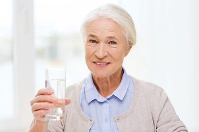 An elderly woman is holding a clear glass of water and smiling at the camera.