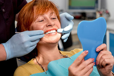 The image shows a woman in a dental chair, smiling and looking at her teeth while holding up a blue oral appliance or mouthguard. She is being attended to by a dentist who is adjusting the appliance with gloved hands.