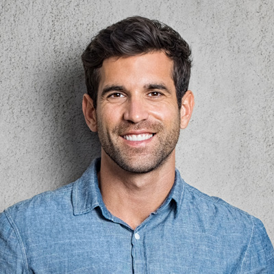 A smiling man with short hair, wearing a blue denim shirt, poses against a concrete background.