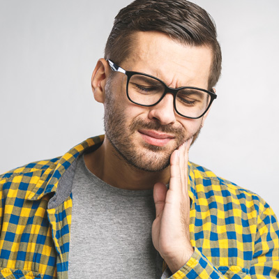 A man with a beard wearing glasses and a plaid shirt, holding his hand to his mouth in pain or discomfort.
