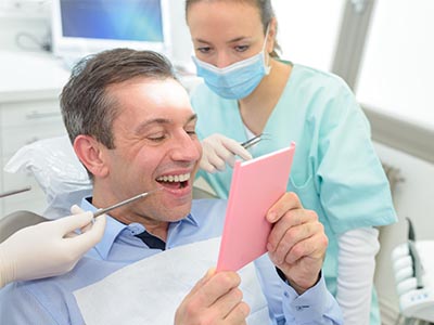 In the image, a man is seated in a dental chair with a pink cardboard sign held up to his face. He appears surprised or amused. Behind him, a female dental professional is smiling and looking at the camera. The setting suggests a dental clinic environment.