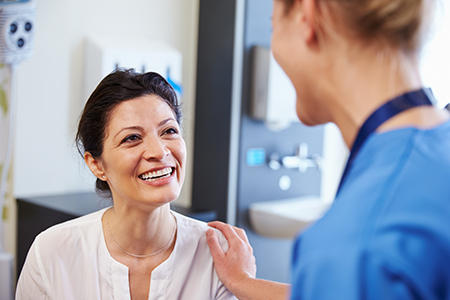 The image is a photograph showing two individuals, likely in a medical setting. A woman with dark hair is smiling at the camera while seated, and behind her stands a person wearing a white lab coat, who appears to be engaged in a conversation or interaction with the woman. Both are indoors, with visible medical equipment and a blue wall in the background.