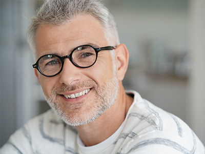 The image features a man with glasses, smiling at the camera. He has gray hair and is wearing a white shirt.