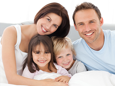 The image shows a family of four, including two adults and two children, posing together in a bedroom setting.