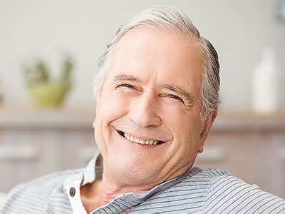 The image shows an elderly man with gray hair, wearing a blue shirt and smiling at the camera. He is seated comfortably in a chair indoors.